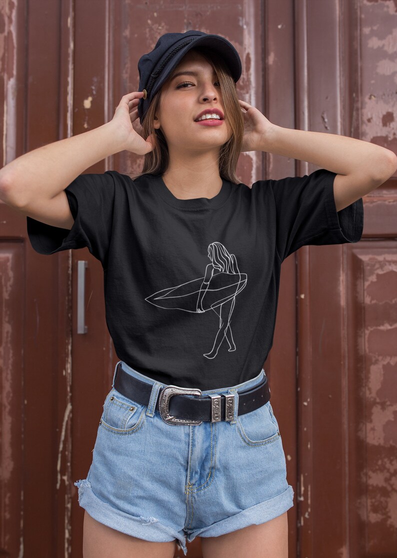 May include: A black t-shirt with a white line drawing of a person surfing. The person is holding a surfboard and walking on the beach. The t-shirt is being worn by a person with brown hair and a black cap.