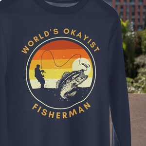 May include: Navy blue long-sleeve sweatshirt with a vintage-style graphic of a fisherman casting a line with a fish jumping out of the water. The graphic is surrounded by a circle with the text "WORLD'S OKAYIST FISHERMAN".