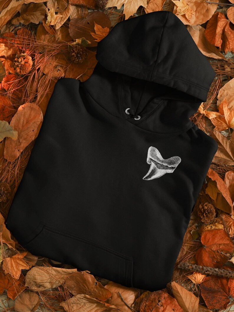 May include: Black hooded sweatshirt with a white embroidered shark tooth design. The sweatshirt is laid out on a bed of brown leaves.