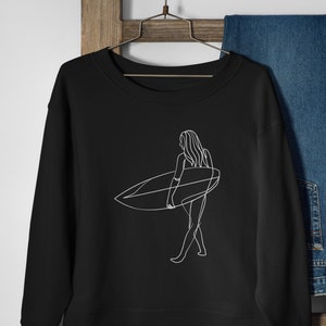 May include: Black sweatshirt with a white line drawing of a person surfing. The person is holding a surfboard and walking on the beach.