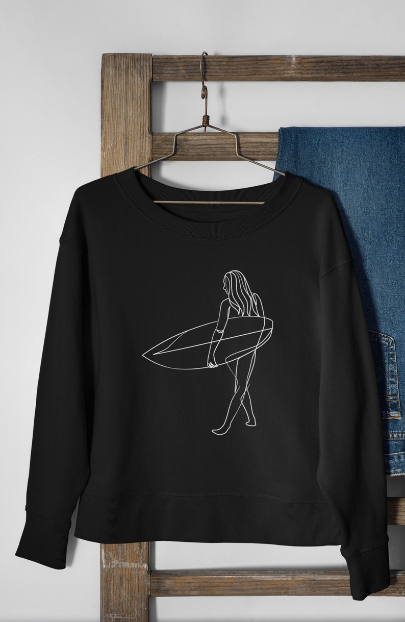 May include: Black sweatshirt with a white line drawing of a person surfing. The person is holding a surfboard and walking on the beach.