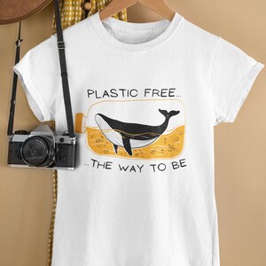 Könnte beinhalten: Weißes T-Shirt mit einem Grafikdruck einer Walin in einer Glasflasche mit dem Text "PLASTIC FREE... THE WAY TO BE"