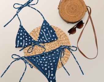 May include: A blue and white patterned bikini with a triangle top and a high-waisted bottom. The bikini is on a woven straw mat with a round straw bag and sunglasses.