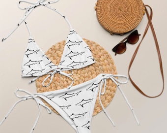 May include: White bikini with a shark print. The bikini top is a triangle style with adjustable straps. The bikini bottom is a tie-side style. The bikini is on a woven straw mat.