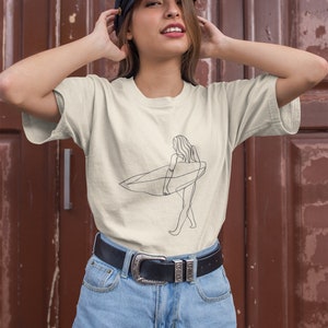 May include: A woman wearing a beige t-shirt with a line drawing of a person surfing. The shirt has a crew neck and short sleeves. The woman is also wearing a black belt with a silver buckle and blue denim shorts.