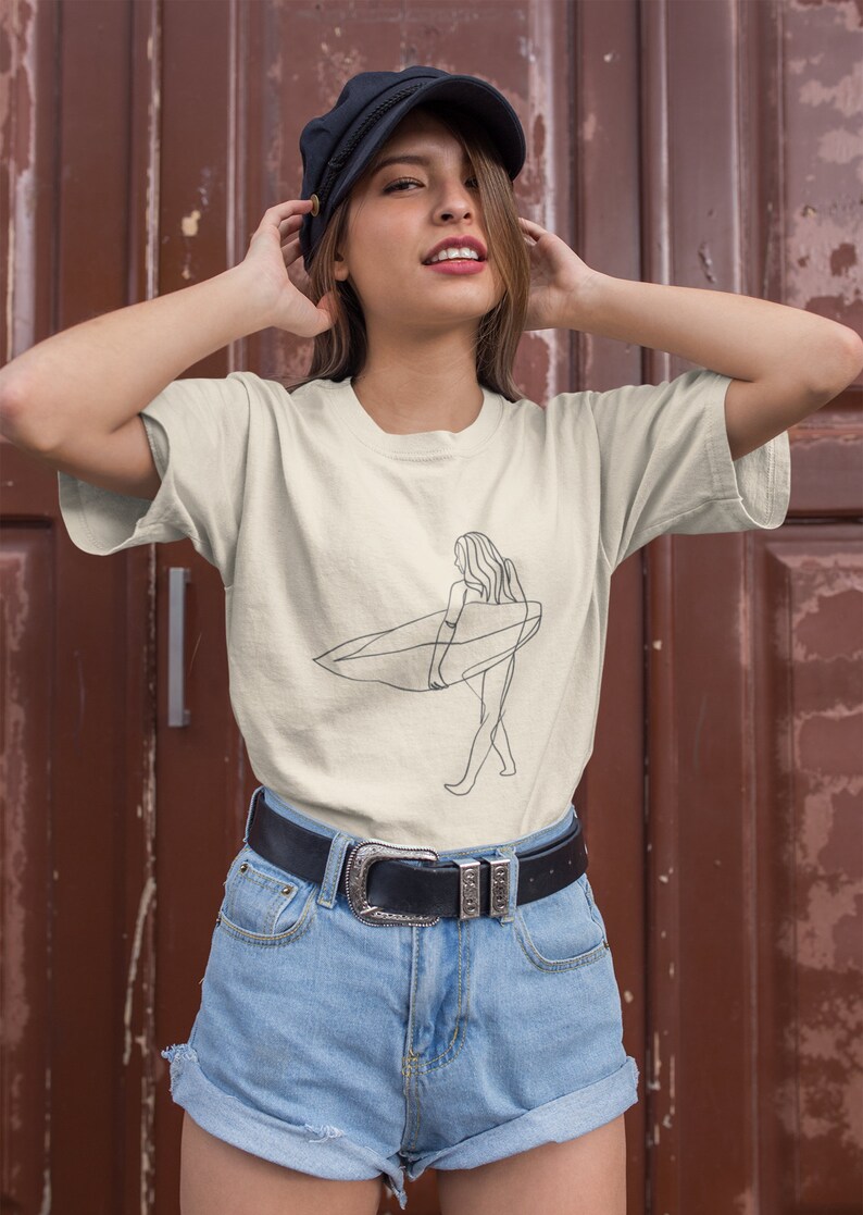 May include: A woman wearing a beige t-shirt with a line drawing of a person surfing. The shirt has a crew neck and short sleeves. The woman is also wearing a black belt with a silver buckle and blue denim shorts.