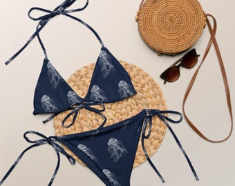 May include: A navy blue bikini with a white jellyfish print. The bikini top is a triangle style with adjustable straps. The bikini bottom is a tie-side style. The bikini is on a woven straw mat.