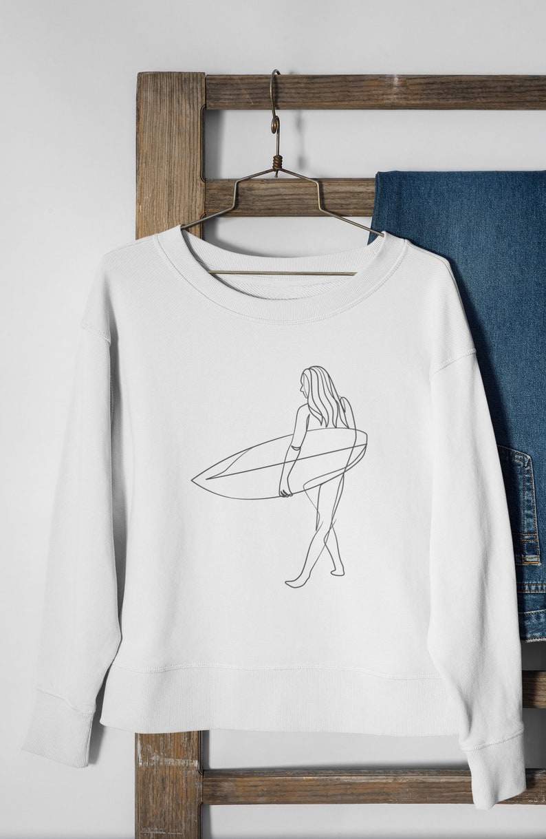 May include: White sweatshirt with a line drawing of a person surfing. The person is holding a surfboard and walking on the beach.