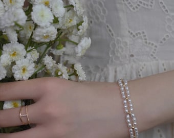 Op de afbeelding: Een dubbele rij parel armband met gouden accenten. De armband zit om de pols van een persoon.
