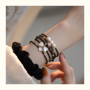 May include: A set of five bracelets featuring black beads, gold accents, and white shell heart charms. One bracelet has three white pearl beads.