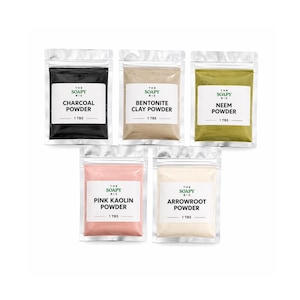 Puede incluir: Cinco bolsas selladas de productos en polvo, cada una etiquetada con el nombre del producto y "1 TBS". Los polvos incluyen carbón vegetal, arcilla bentonita, neem, caolín rosa y arrurruz. Las bolsas están dispuestas sobre un fondo blanco.