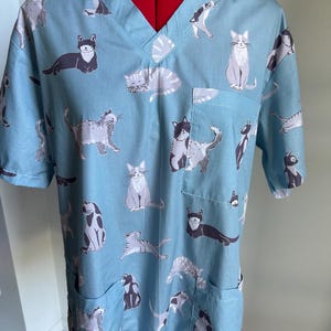 May include: Pale blue scrub top with a V-neck and short sleeves. The top is decorated with grey and black cat designs. It has two front pockets. A practical garment for medical staff or cat enthusiasts.