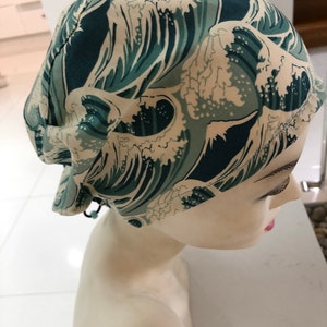 May include: A fabric cap with a teal and cream wave pattern, worn on a mannequin head. The cap has a gathered design and a drawstring. The wave pattern is reminiscent of Japanese art.