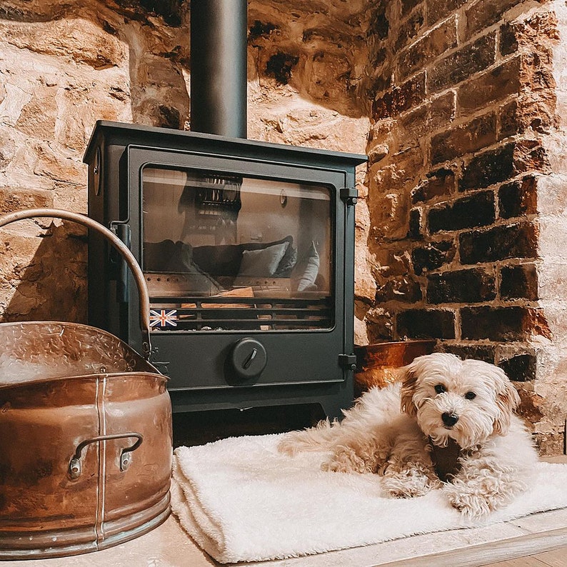 May include: A white fluffy dog sits in front of a black wood-burning stove with a copper bucket and a white rug. The stove is set against a brick wall.