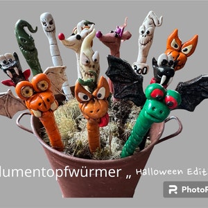 Op de afbeelding: Een verzameling Halloween-thema keramische wormvormige potten in een bruine metalen emmer. De wormen zijn versierd met verschillende griezelige gezichten en kenmerken, waaronder een geest, een vleermuis, een schedel en een draak.