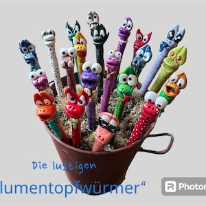 Op de afbeelding: Een verzameling grillige, kleurrijke wormvormige beeldjes met expressieve gezichten, tentoongesteld in een kleine, roestige metalen emmer. De wormen zijn geschilderd in verschillende kleuren, waaronder rood, blauw, groen en paars, met witte ogen en unieke gelaatstrekken.