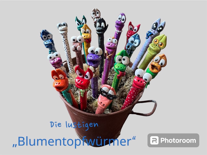 May include: A collection of whimsical, colorful worm-shaped figurines with expressive faces, displayed in a small, rusty metal bucket. The worms are painted in various colors, including red, blue, green, and purple, with white eyes and unique facial features.