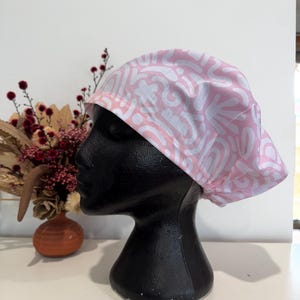 May include: Pink scrub hat with a white abstract pattern. The hat is made of soft, breathable fabric and is ideal for nurses, doctors, and other healthcare professionals.