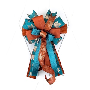 May include: A large, teal and rust-colored bow with gold leaf accents. The bow is made of wired ribbon and is tied in a classic, symmetrical shape.