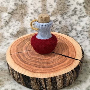 May include: A crocheted potion bottle with a red body, light blue neck, and a tan cork. The bottle is sitting on a wooden slice.
