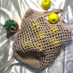 Peut inclure: Un sac en filet beige crocheté rempli de pommes jaunes. Le sac a une base rectangulaire et deux anses. Une petite plante succulente en pot et deux pommes supplémentaires sont également visibles.