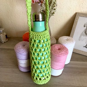Peut inclure: Un porte-bouteille d'eau crocheté vert clair avec une sangle assortie, contenant une bouteille d'eau vert menthe. Plusieurs bobines de fil de différentes couleurs, dont lavande, rose, orange et blanc, sont disposées autour de la bouteille.
