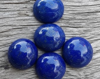 May include: Five polished, round, deep blue lapis lazuli stones. The stones have a smooth, glossy surface with flecks of gold. The stones are arranged on a weathered, gray wooden surface.