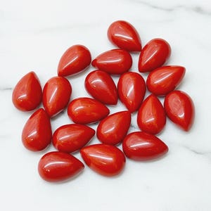 May include: A collection of polished, teardrop-shaped red gemstones. The stones have a glossy finish and are scattered across a white surface. The gemstones are a vibrant, solid red color.