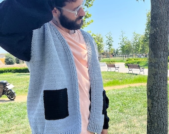 May include: A light blue crocheted cardigan with black sleeves and a black patch pocket. The cardigan is open in the front and has a relaxed fit.
