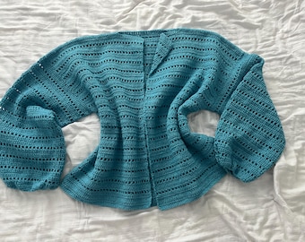 May include: A teal blue crocheted cardigan with a loose fit and long sleeves. The cardigan has a V-neck and a delicate open stitch pattern.