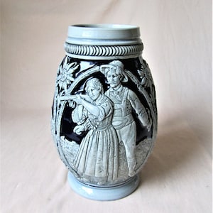 Könnte beinhalten: Ein blau-weißer Bierkrug aus Keramik mit einem detaillierten Relief eines Mannes und einer Frau in Tracht, die ein Gewehr anvisieren. Der Krug hat florale und dekorative Akzente.