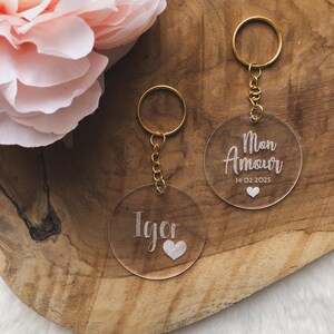 Op de afbeelding: Twee transparante acryl sleutelhangers met gouden sleutelringen. Eén sleutelhanger heeft het woord "Igor" en een hart gegraveerd. De andere sleutelhanger heeft de woorden "Mon Amour" en de datum "14 02 2025" gegraveerd.