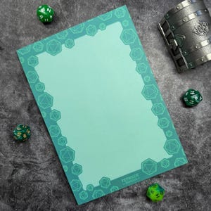 May include: A teal notepad with a border of geometric shapes, possibly for tabletop gaming. The notepad has the text "Feywild Supply Co." at the bottom right. Several green dice and a metal treasure chest are scattered around the notepad.