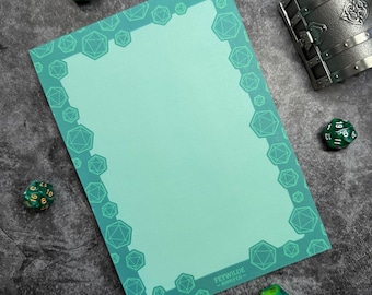 May include: A teal-colored notepad with a border of geometric shapes, likely for tabletop gaming. The notepad has the text "Feywild Supply Co." at the bottom right. Several green dice and a metal treasure chest are scattered around the notepad.