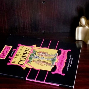 Può includere: Un libro intitolato "MORIRE di COPPIA" con copertina nera e dettagli rosa e gialli. Il libro è su una superficie di legno scuro, con una scultura astratta dorata sullo sfondo.