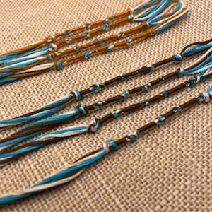 Peut inclure: Un ensemble de quatre bracelets d'amitié en fils bleus, blancs et bruns. Les bracelets sont noués ensemble et ont un pompon à l'extrémité.