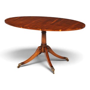 Può includere: Un tavolo ovale in legno con una ricca venatura marrone, sostenuto da una colonna centrale che si dirama in quattro gambe curve. Ogni gamba termina con una piccola rotella in ottone, fornendo una base stabile per la superficie del tavolo.