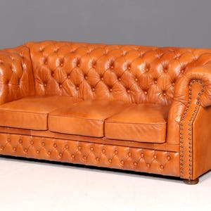 Könnte beinhalten: Ein dreisitziges, getuftetes Ledersofa in einem warmen Karamellton. Das Sofa hat gerollte Armlehnen, Knopfdetails und Ziernagelbesatz an der Vorderseite und den Armlehnen. Ein klassisches Möbelstück.