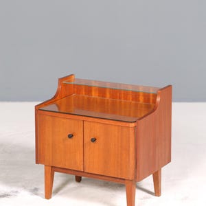 Può includere: Un mobile in legno in stile mid-century moderno, con due ante e un piano in vetro. Il mobile è di colore marrone caldo, con pomelli neri e gambe affusolate. Un design semplice ed elegante per una camera da letto o un soggiorno.