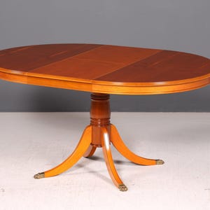 Può includere: Un tavolo da pranzo ovale in legno con una ricca finitura marrone sul piano e una base in legno color miele più chiaro. Il tavolo presenta una colonna centrale a piedistallo che si dirama in quattro gambe curve, ciascuna terminante con un piede in metallo color ottone.