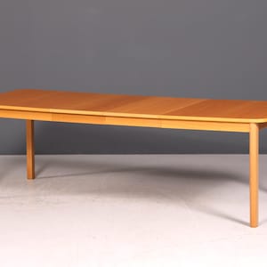 Peut inclure: Table de salle à manger en bois brun clair avec quatre pieds droits. Le plateau rectangulaire a une finition brun clair et semble avoir une rallonge à feuilles. La table est placée sur un fond gris.