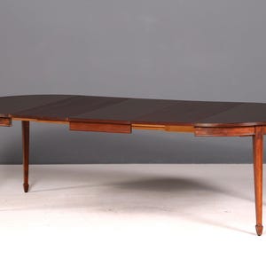 Peut inclure: Table de salle à manger ovale en bois avec un plateau marron foncé et quatre pieds effilés. La table a une fonction d'extension, avec une section qui peut être retirée pour augmenter la surface. Le bois a une teinte brun rougeâtre chaude.