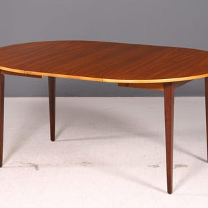 Peut inclure: Une table à manger en bois de forme ovale avec une surface au grain brun riche et une bordure en bois plus clair. La table est soutenue par quatre pieds fuselés et fins qui s'étendent vers l'extérieur depuis le centre de la structure.