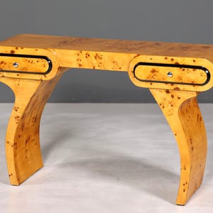 Könnte beinhalten: Eine hellfarbene Holzkonsolentisch mit zwei Schubladen und geschwungenen Beinen. Die Schubladen haben schwarze Zierleisten und silberne Beschläge. Das Holz hat ein gesprenkeltes Muster.
