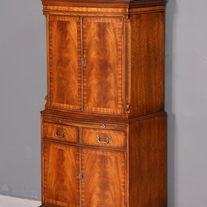 Può includere: Un mobile alto antico in legno con una ricca finitura marrone-rossastra. Il mobile presenta due ante superiori, due cassetti e due ante inferiori, tutti con ferramenta ornata. Il design include modanature decorative e una base curva.