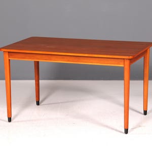 Peut inclure: Une table rectangulaire en bois avec une finition brun moyen lisse. La table présente quatre pieds droits et effilés, chacun se terminant par un petit embout cylindrique noir. Le plateau a des coins arrondis et une surface plane et épurée.