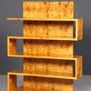 Op de afbeelding: Een lichtgekleurde houten boekenplank met een uniek, zig-zag ontwerp. De planken zijn rechthoekig en open, met een natuurlijk houtnerfpatroon. De boekenplank heeft een verticale achterkant en een stevige basis.