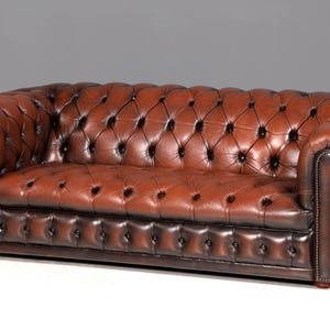Puede incluir: Un sofá Chesterfield de cuero marrón con diseño capitoné. El sofá tiene brazos enrollados y un perfil bajo. El cuero tiene un acabado brillante y el sofá está sobre un fondo neutro. Mide aproximadamente 200 cm de largo.
