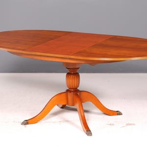 Può includere: Un tavolo da pranzo ovale in legno con una finitura marrone calda e una superficie liscia. Il tavolo presenta una base a piedistallo centrale con una colonna scanalata e quattro gambe curve che terminano con piedi metallici a forma di artiglio.
