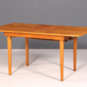 Op de afbeelding: Een lichtbruine houten eettafel met vier taps toelopende poten. Het rechthoekige tafelblad heeft een lichtere houtnerf en lijkt een bladverlenging te hebben. De tafel staat tegen een grijze achtergrond.